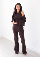 A woman modeling a dark brown knit lounge set consisting of a cold-shoulder long-sleeve top and matching wide-leg pants, standing in a bright, minimalist room with white walls and light wood flooring.