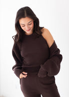 A woman wearing a dark brown ribbed knit sweater with an asymmetrical off-the-shoulder design and matching dark brown knit pants, photographed against a clean white studio background.