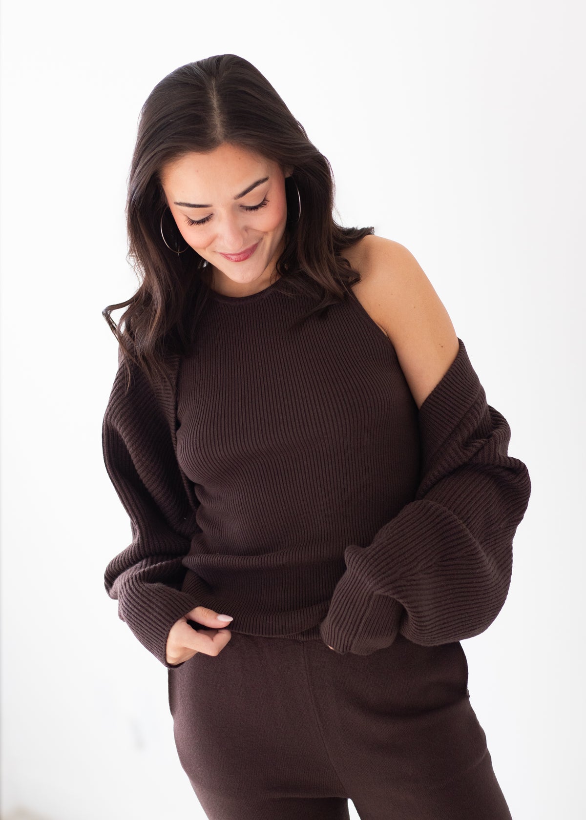 A woman wearing a dark brown ribbed knit sweater with an asymmetrical off-the-shoulder design and matching dark brown knit pants, photographed against a clean white studio background.