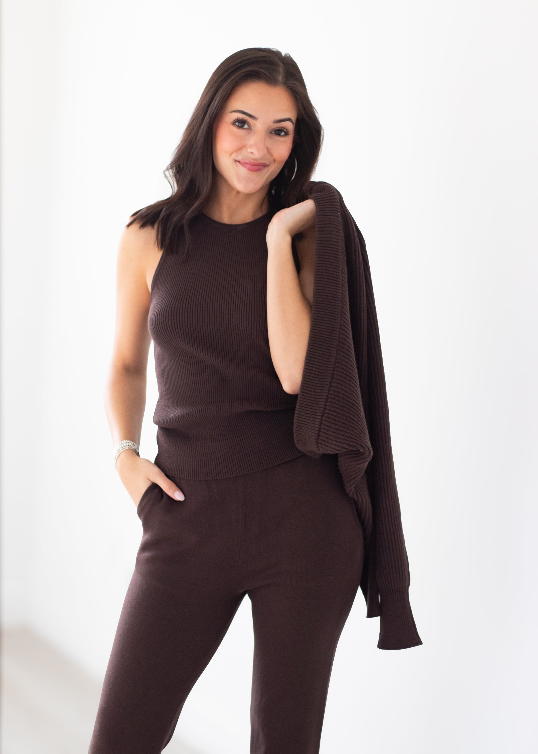 A woman modeling a dark brown ribbed knit set consisting of a sleeveless top and matching pants, with a matching knit cardigan draped over her shoulder, against a plain white studio background.