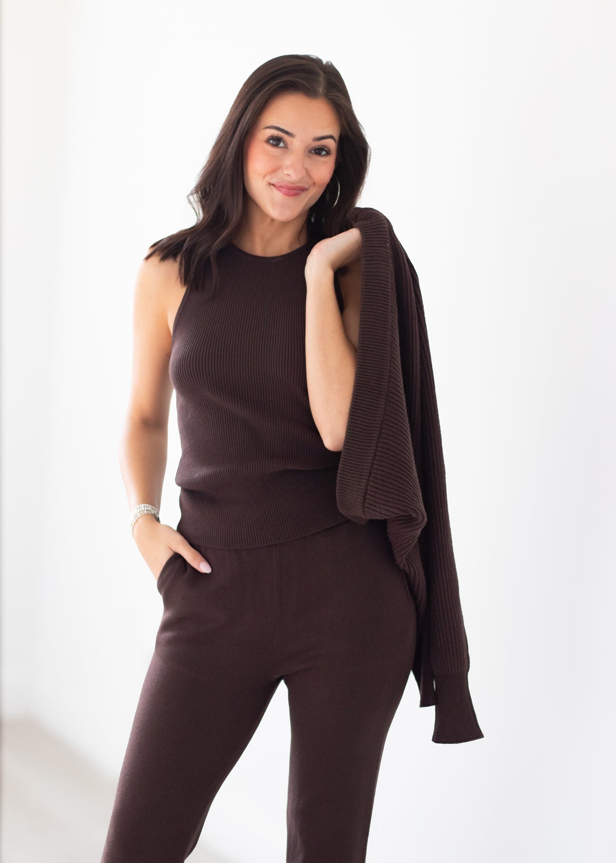 A woman modeling a dark brown ribbed knit set consisting of a sleeveless top and matching pants, with a matching knit cardigan draped over her shoulder, against a plain white studio background.