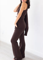 A woman modeling a dark brown ribbed knit jumpsuit with a sleeveless bodice, side pockets, and wide-leg pants, standing against a bright white wall.