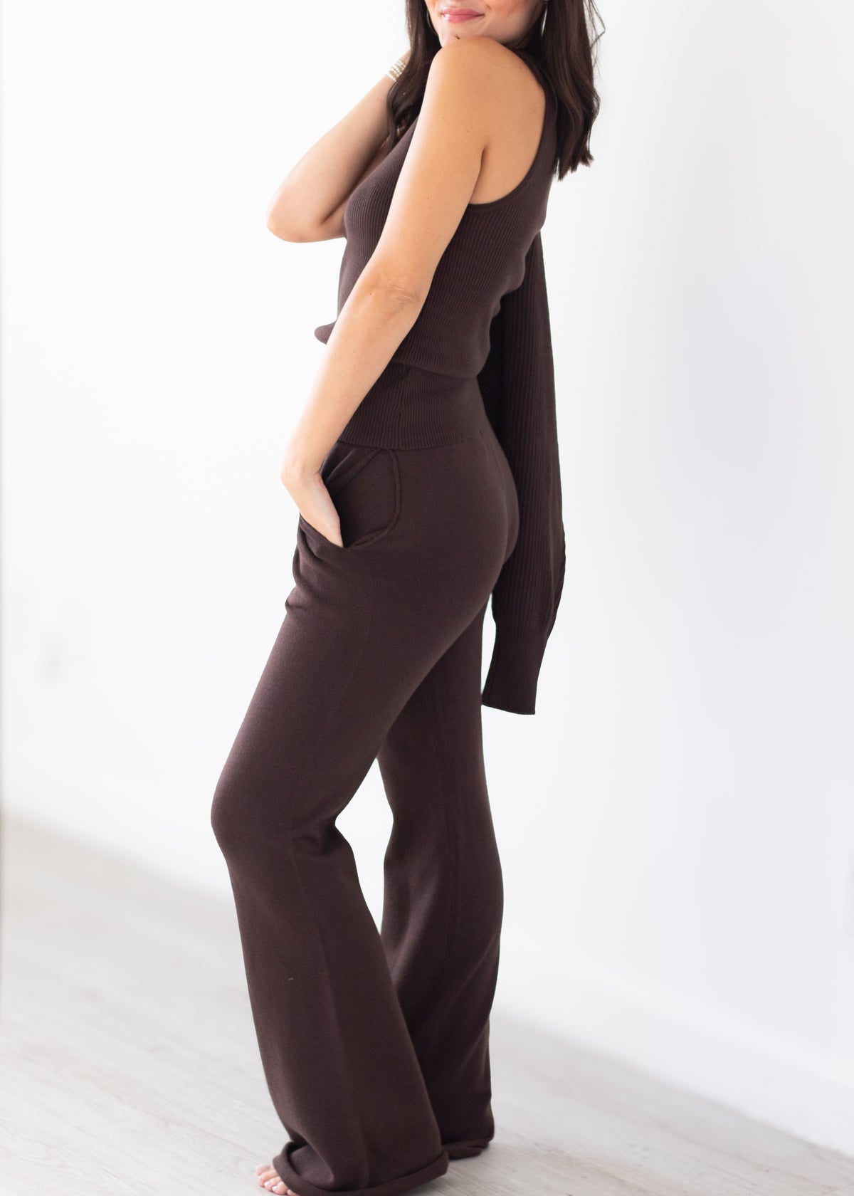 A woman modeling a dark brown ribbed knit jumpsuit with a sleeveless bodice, side pockets, and wide-leg pants, standing against a bright white wall.