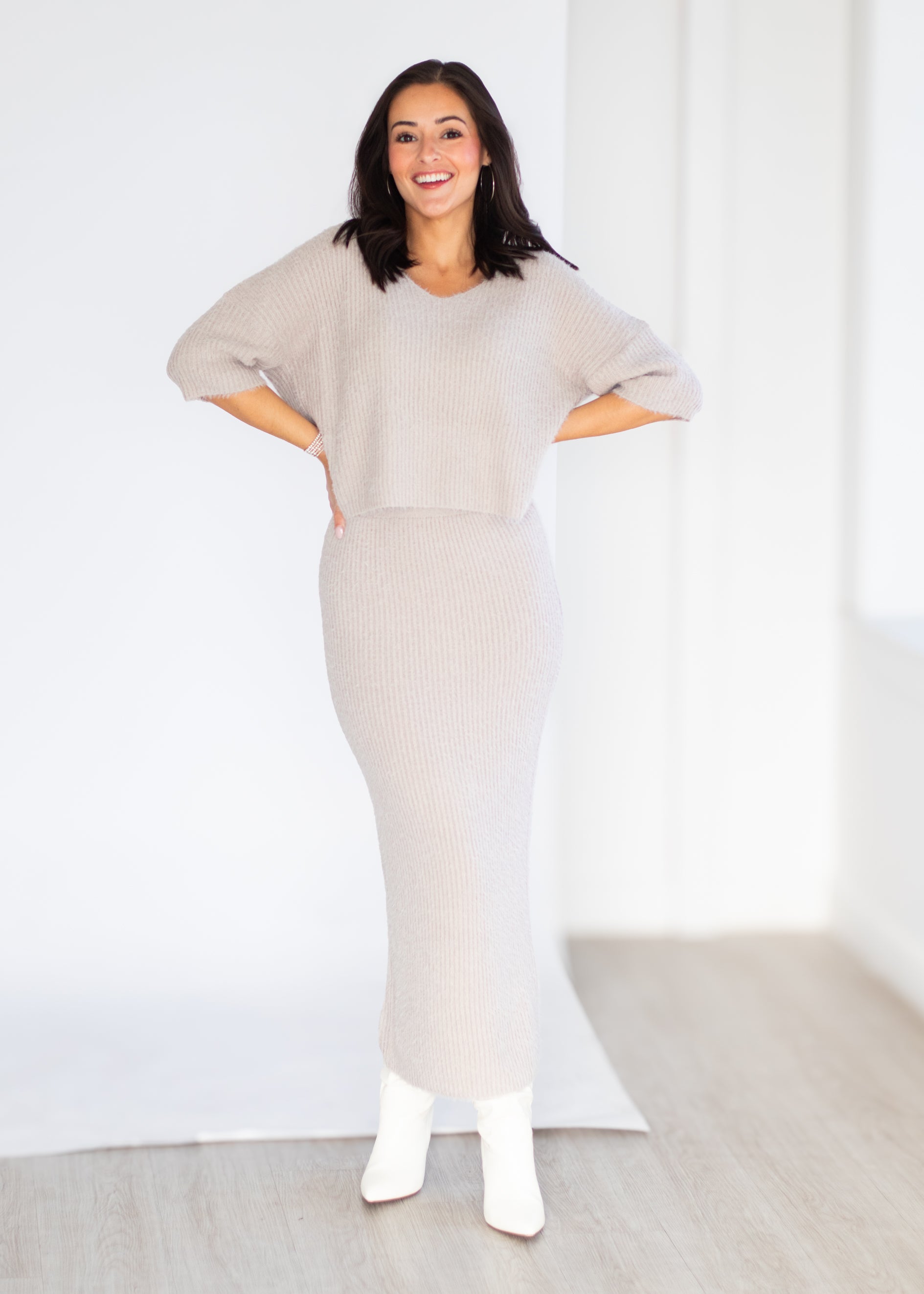 A woman modeling a light beige ribbed knit set consisting of a short-sleeve cropped sweater and a matching ribbed midi skirt, paired with white ankle boots, in a bright studio setting.