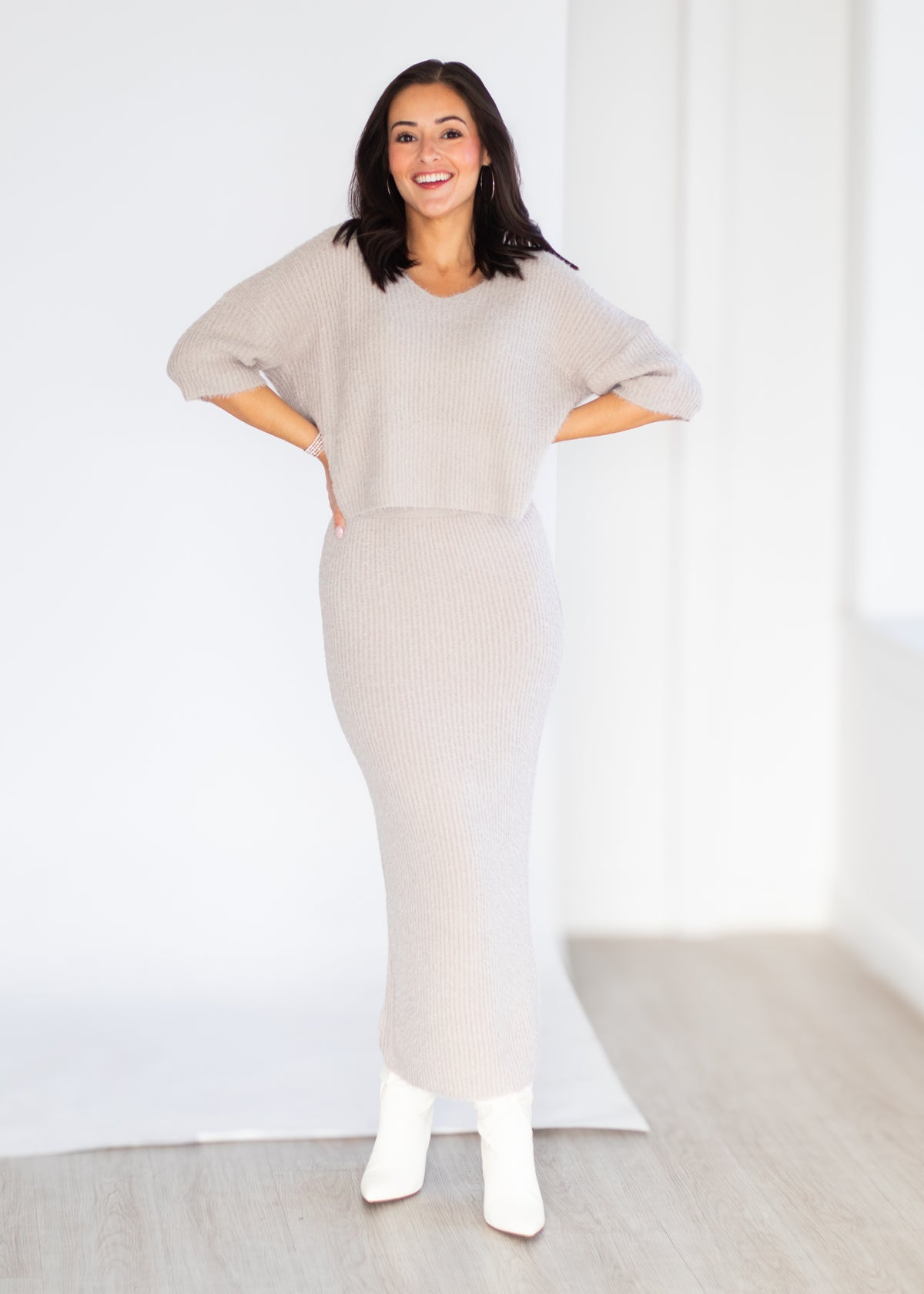 A woman modeling a light beige ribbed knit set consisting of a short-sleeve cropped sweater and a matching ribbed midi skirt, paired with white ankle boots, in a bright studio setting.