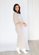 A woman modeling a light beige ribbed knit maxi dress with elbow-length sleeves, paired with white heeled ankle boots, standing against a white studio backdrop.