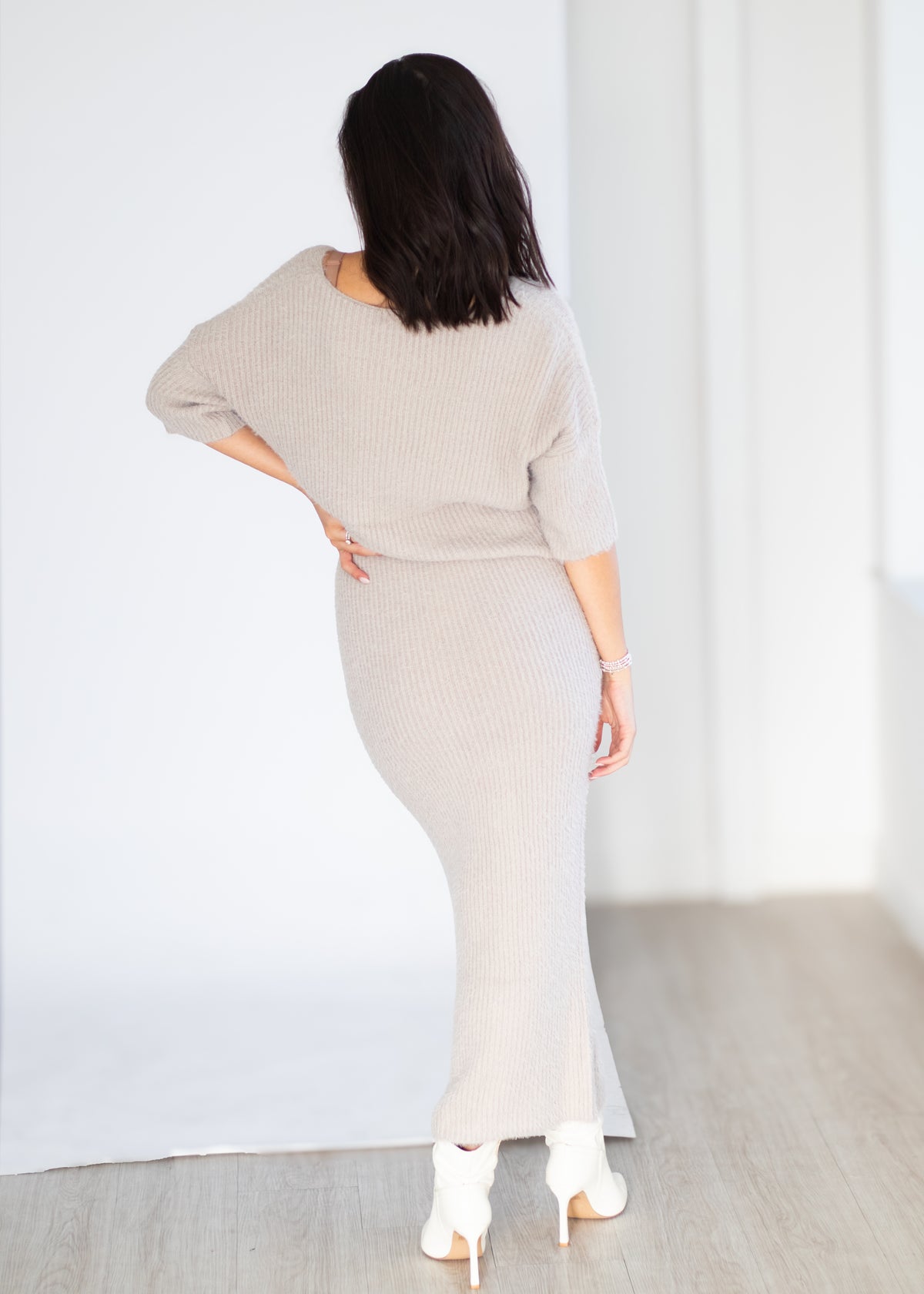 A woman shown from the back wearing a light beige ribbed knit two-piece set consisting of a wide three-quarter sleeve top and a matching long ribbed skirt, paired with white pointed-toe heels.