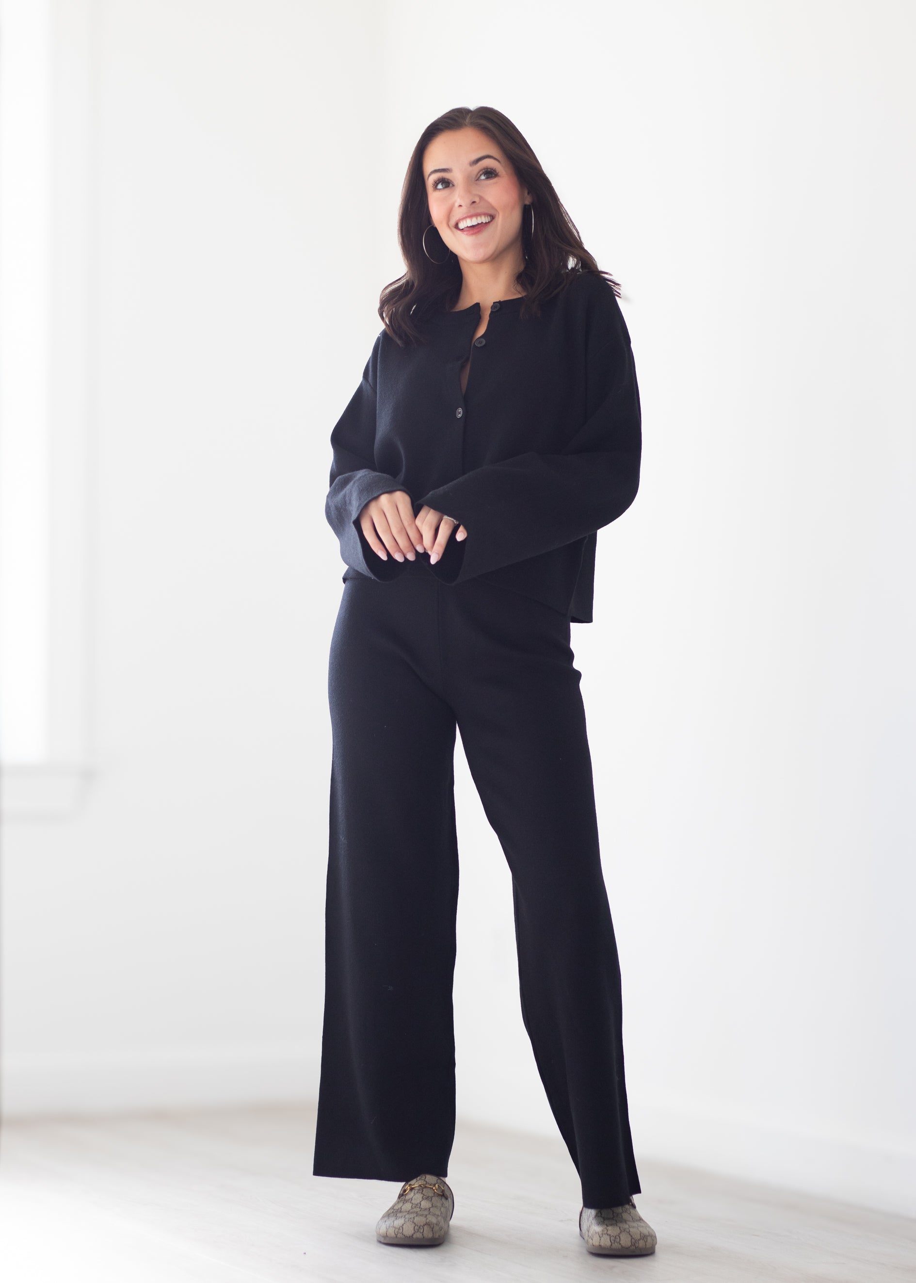 A woman modeling a black long-sleeve button-front top and matching black wide-leg pants in a bright, minimalist white room.