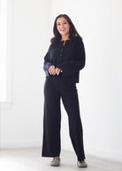 A woman modeling a black long-sleeve button-front top and matching black wide-leg pants in a bright, minimalist white room.