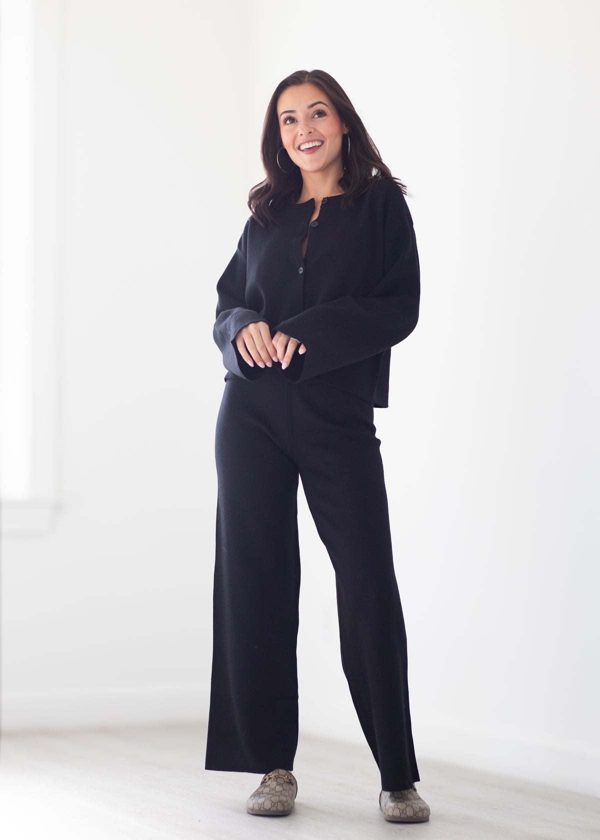 A woman modeling a black long-sleeve button-front top and matching black wide-leg pants in a bright, minimalist white room.
