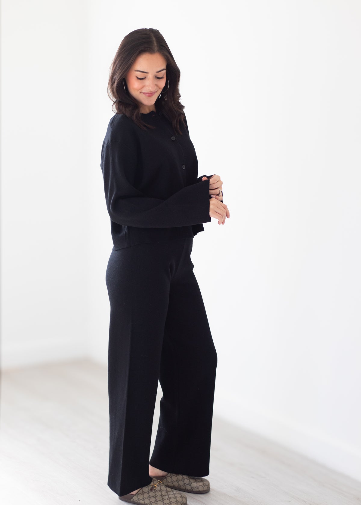 A woman modeling a black long-sleeve button-front top and matching black wide-leg pants, standing in a bright white studio with light wood flooring.