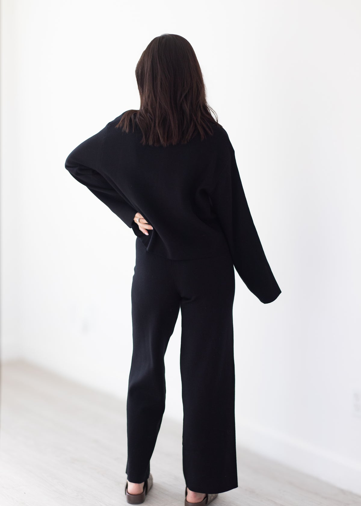 A woman viewed from behind wearing a black knit two-piece outfit consisting of a long-sleeve cropped sweater and matching wide-leg pants, standing in a minimalist room with a white wall and light wood floor.
