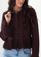 A woman wearing a dark brown textured knit cardigan with fuzzy fringe trim on the sleeves and hem, paired with light blue jeans against a plain white background.