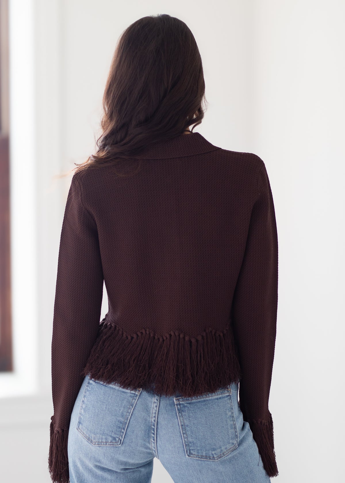 A woman viewed from behind wearing a dark brown ribbed knit cardigan with fringe detailing at the hem and cuffs, paired with light blue jeans.