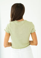 Woman wearing a green short-sleeve cardigan and blue jeans against a neutral background

