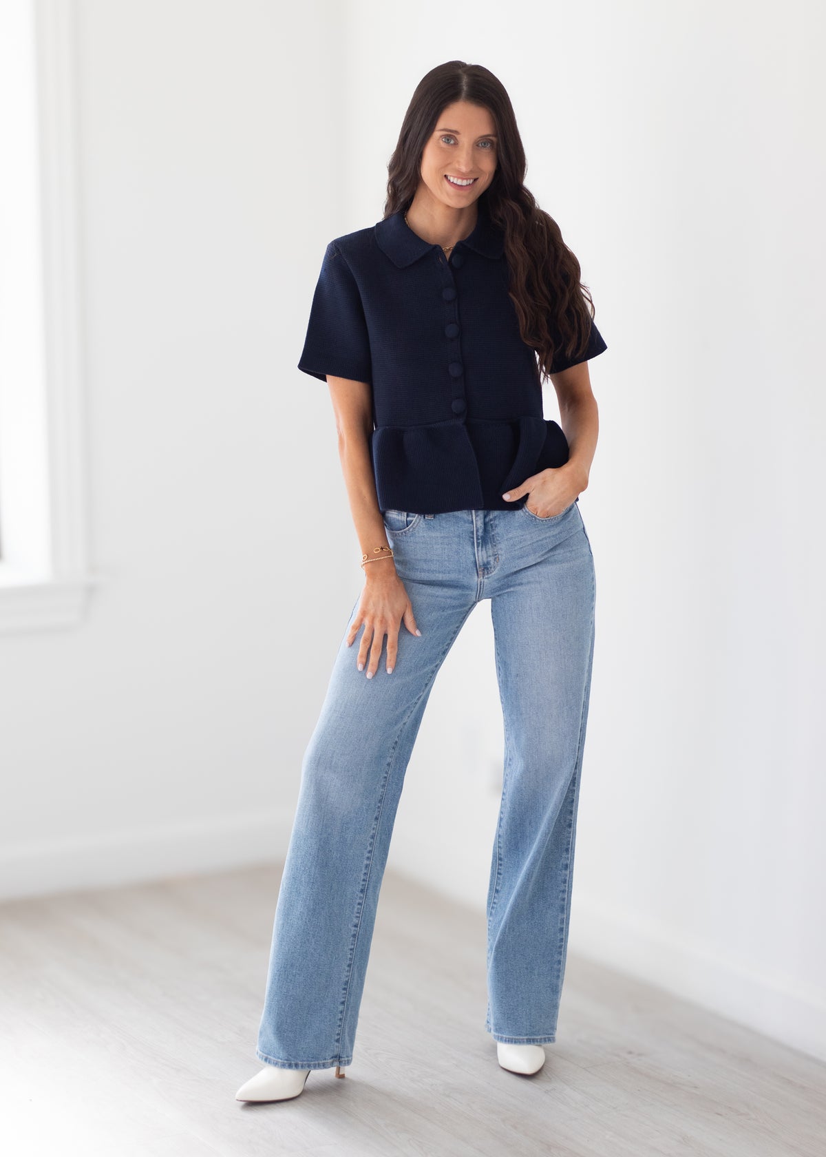 A woman modeling a short-sleeve dark navy button-front top with a peplum waist, paired with light-wash wide-leg jeans and white pointed shoes, standing in a bright white room.