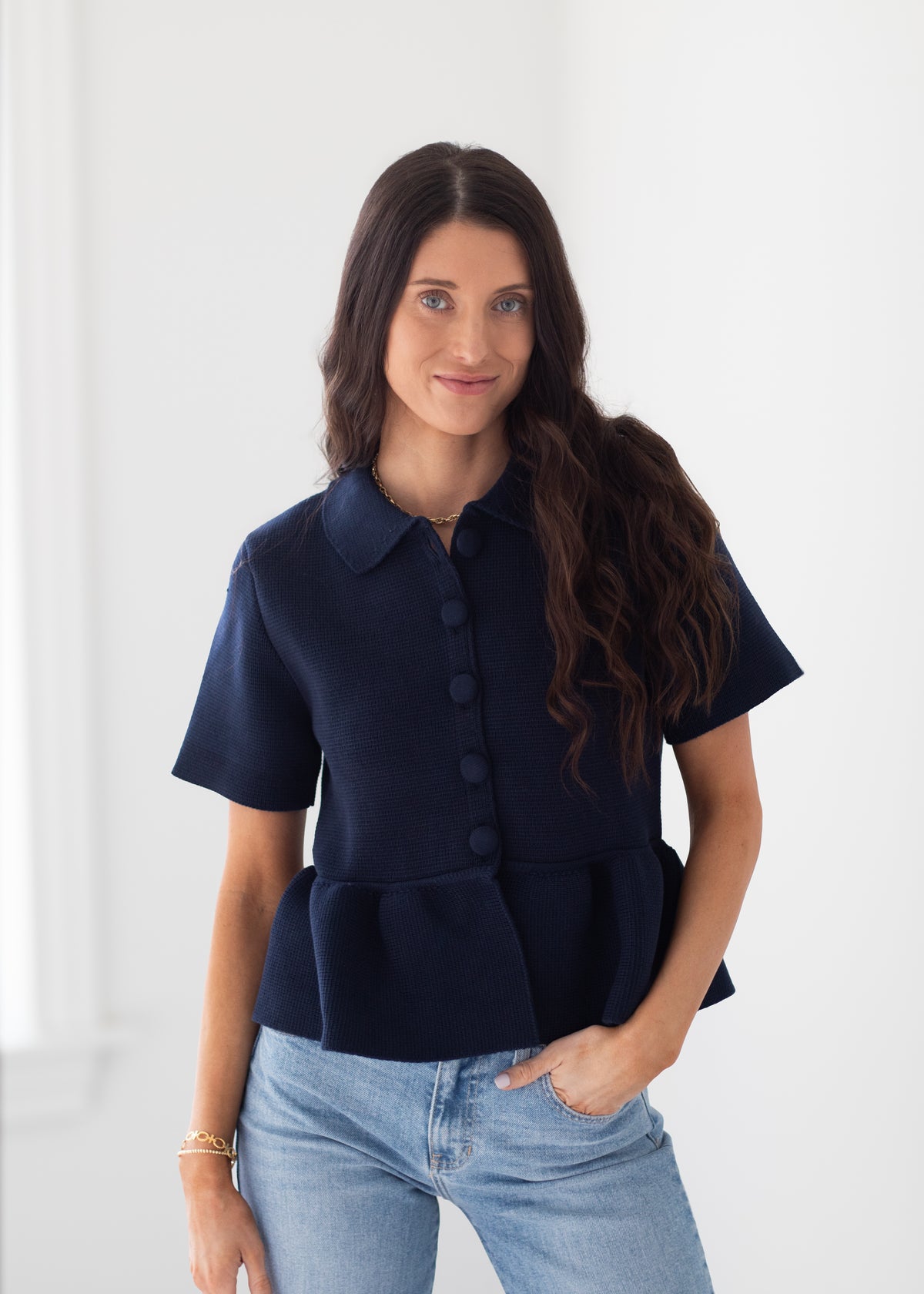 A woman modeling a navy blue short-sleeve button-up peplum top with a pointed collar, paired with light blue jeans, set against a bright white indoor background.