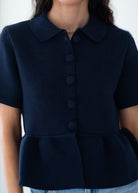 A close-up of a model wearing a navy blue short-sleeve knit top with a collar, button-front closure, and a peplum hem.
