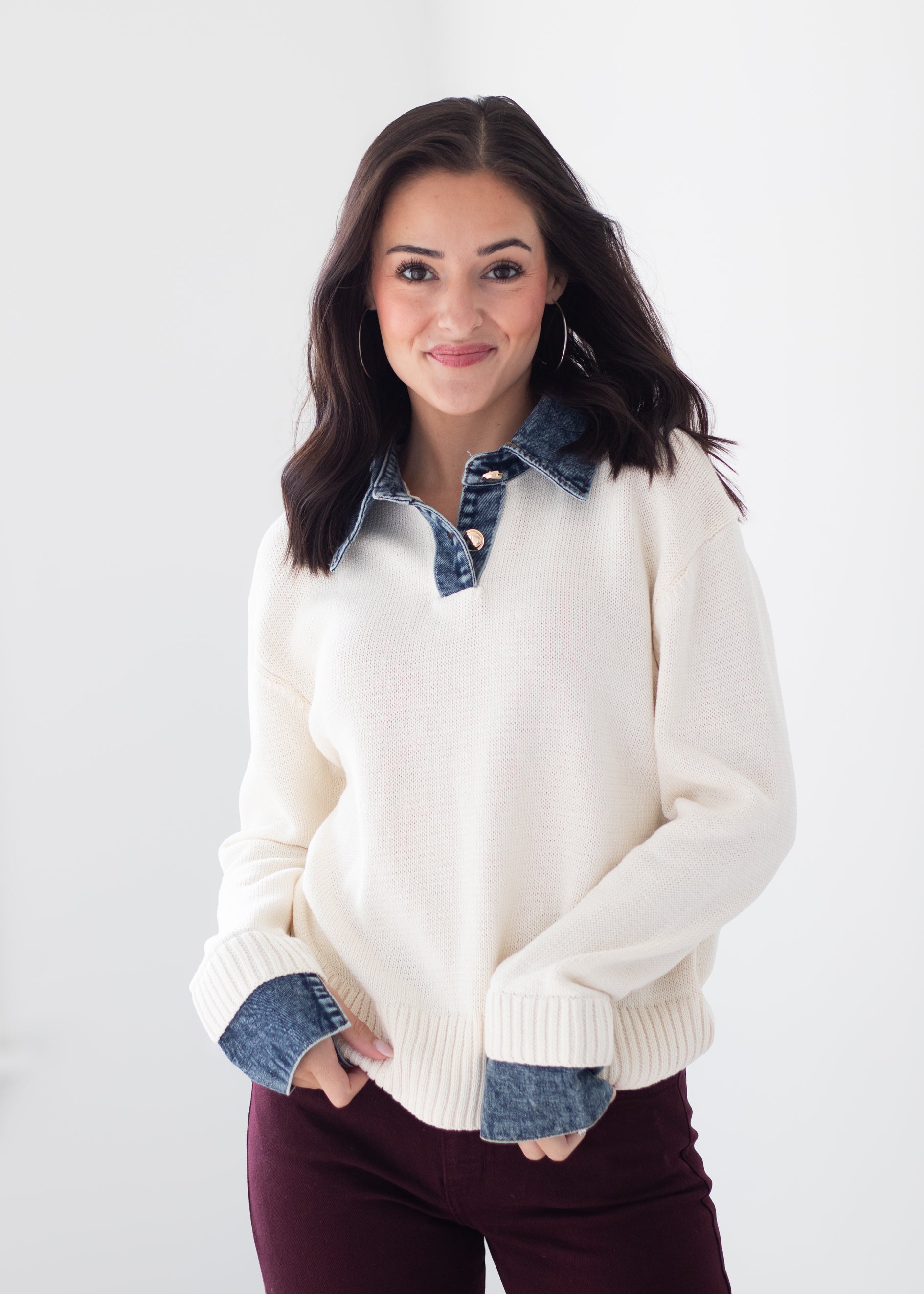 A woman modeling a cream knit sweater with contrasting blue denim collar and cuff details, paired with dark burgundy pants, against a plain white studio background.