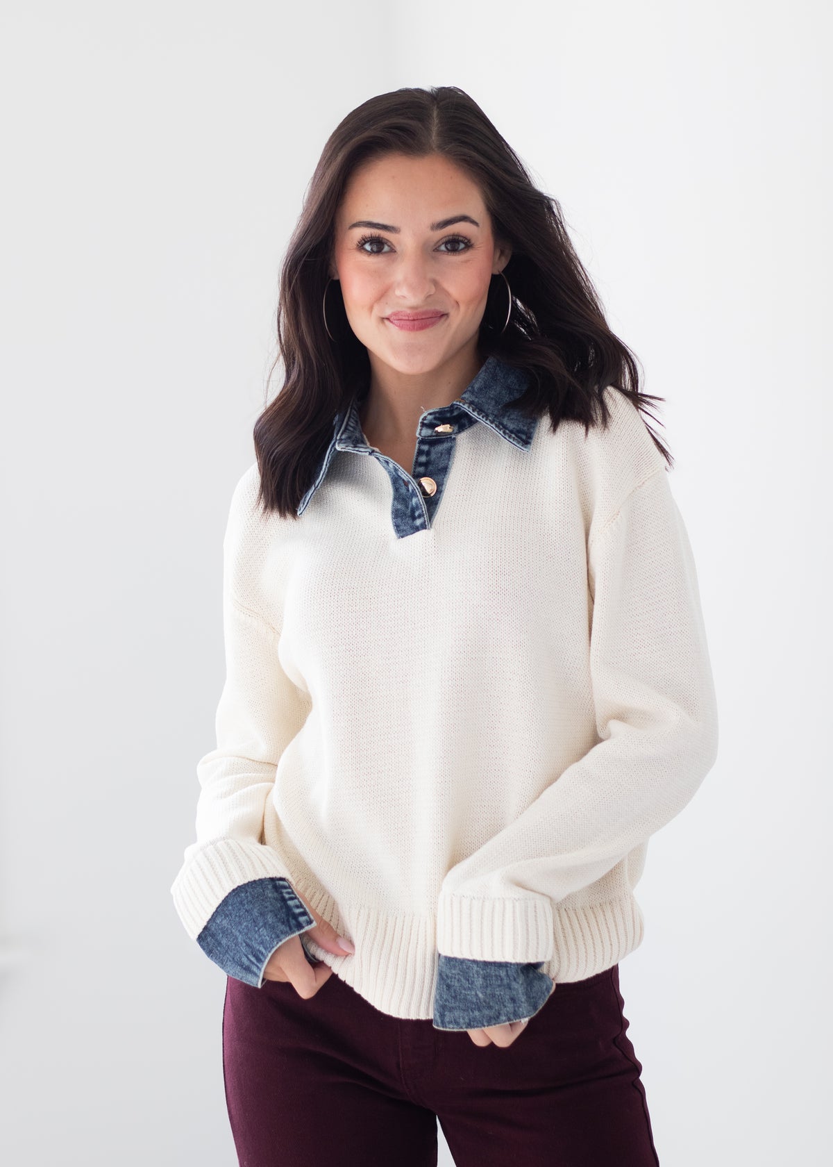 A woman modeling a cream knit sweater with contrasting blue denim collar and cuff details, paired with dark burgundy pants, against a plain white studio background.