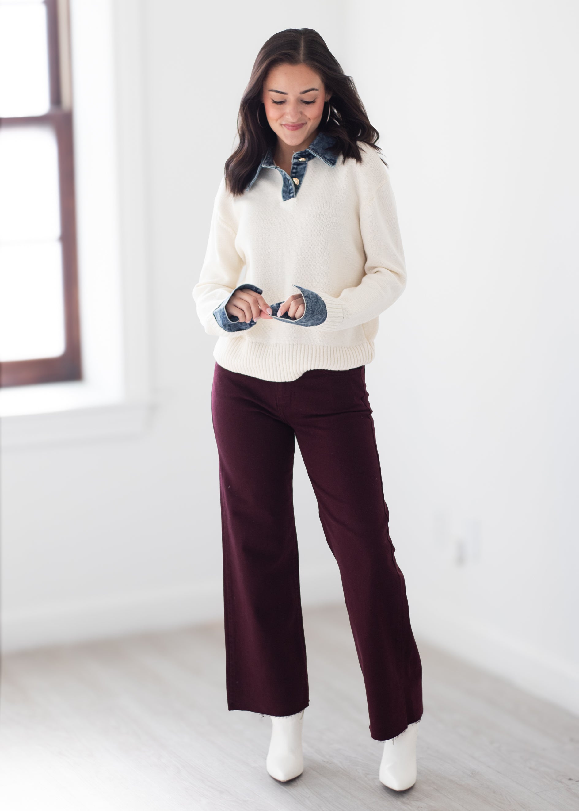 A woman modeling a cream ribbed knit sweater with a dark blue denim collar and cuffs, paired with dark burgundy wide-leg pants and white ankle boots, standing in a bright white room.