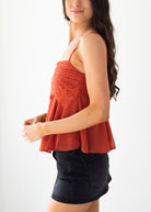 A woman wearing a rust-orange camisole top with delicate lace embroidery on the bodice and a flared peplum hem, paired with a black denim mini skirt, shown from a side profile against a plain white background.