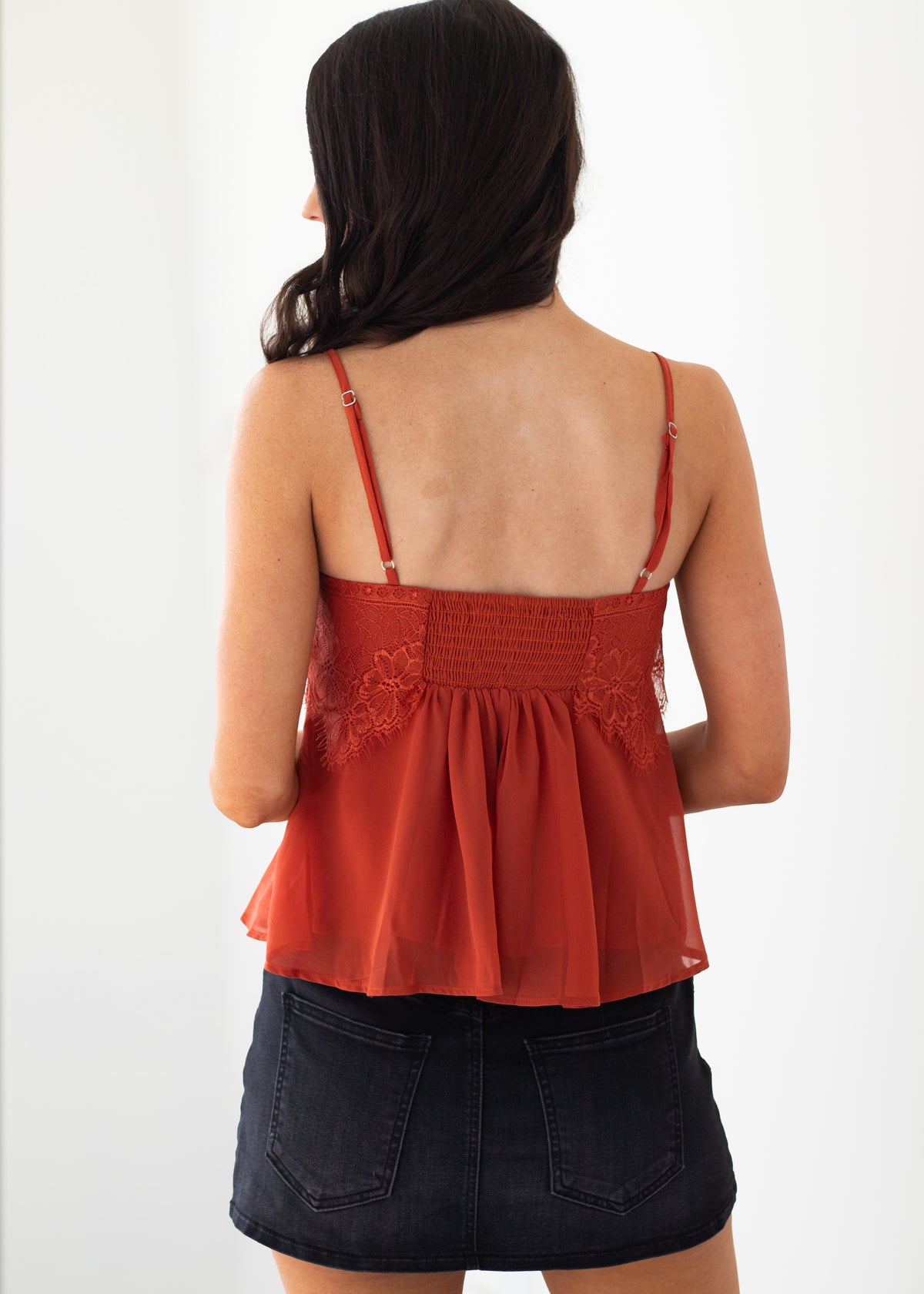 A woman shown from the back wearing a rust-orange spaghetti strap camisole top with lace and smocked detailing, paired with a dark denim mini skirt against a plain white background.