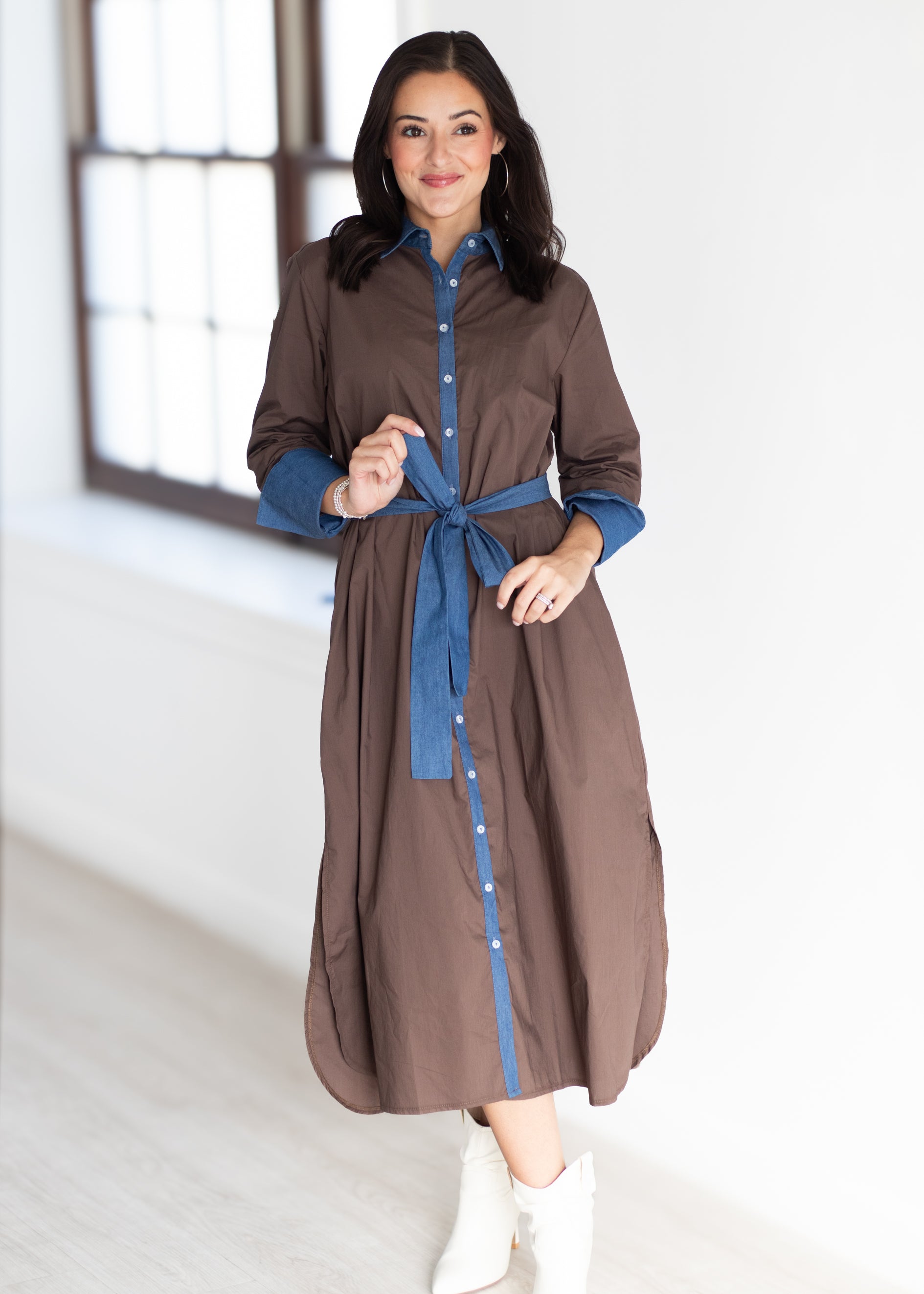 A woman modeling a brown button-front shirt dress with blue shirt-sleeves and a matching blue tie belt, paired with white ankle boots in a bright indoor room.