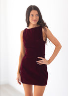 A woman modeling a sleeveless, off-the-shoulder mini dress in deep burgundy velvet against a bright white studio background.