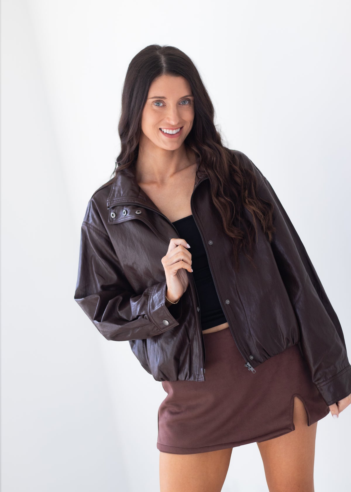 A woman modeling a dark brown faux-leather jacket over a black crop top and a matching brown mini skirt, set against a plain white studio background.