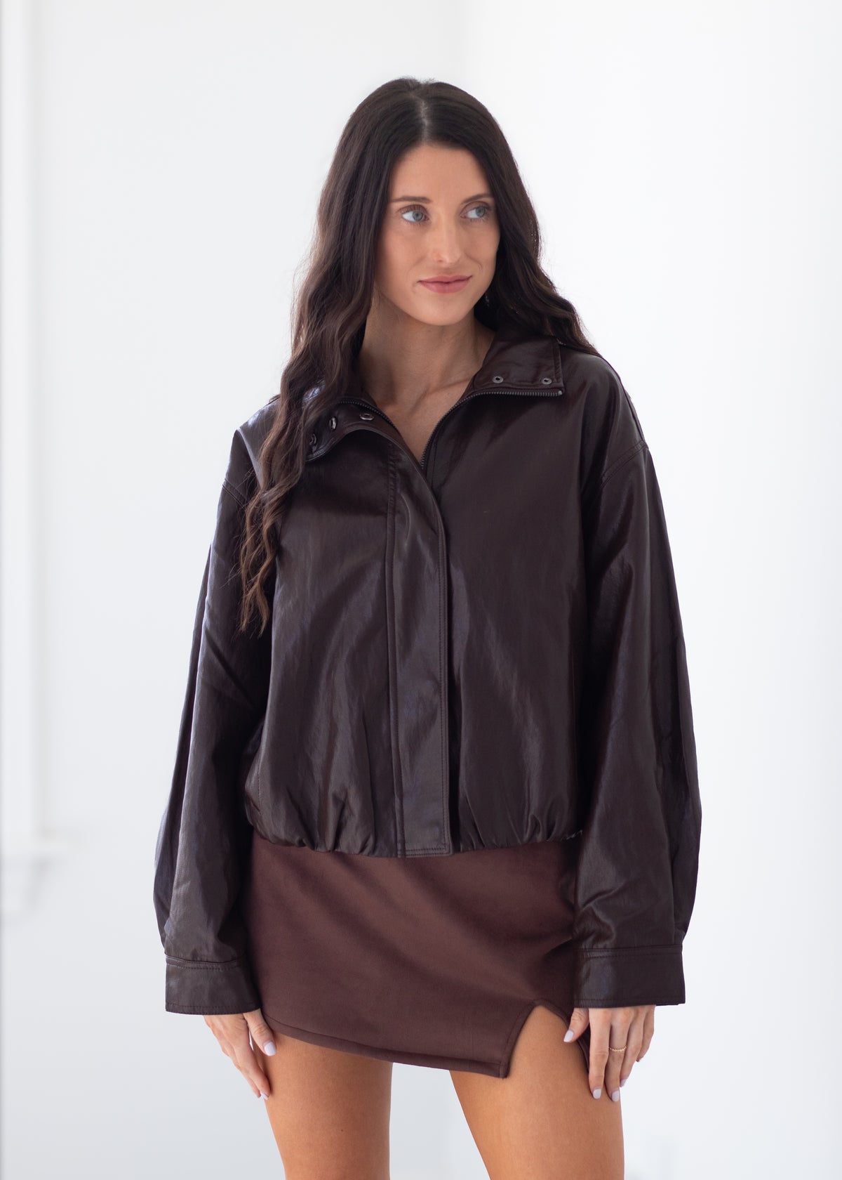 A woman modeling a dark brown faux-leather jacket with a high collar and button front, paired with a matching brown mini skirt, against a plain white studio background.