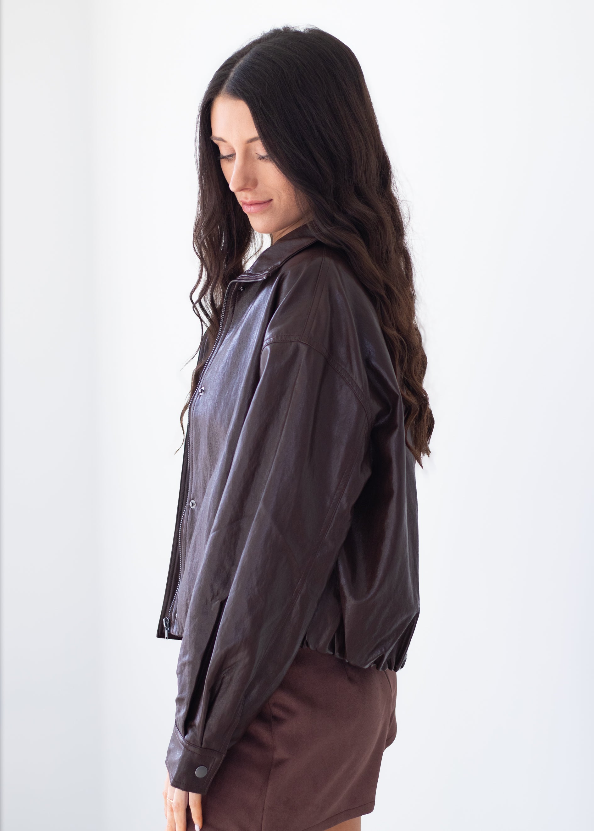 A woman with long dark wavy hair is shown in profile wearing a dark brown, semi-sheer lightweight jacket over a white top and a brown skirt, set against a plain white background.