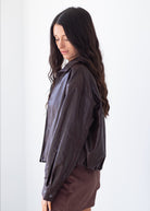 A woman with long dark wavy hair is shown in profile wearing a dark brown, semi-sheer lightweight jacket over a white top and a brown skirt, set against a plain white background.