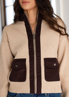 A woman wearing a beige knit zip-up cardigan with dark brown contrast panels, including a hood, zipper trim, and two front flap pockets, paired with blue jeans.