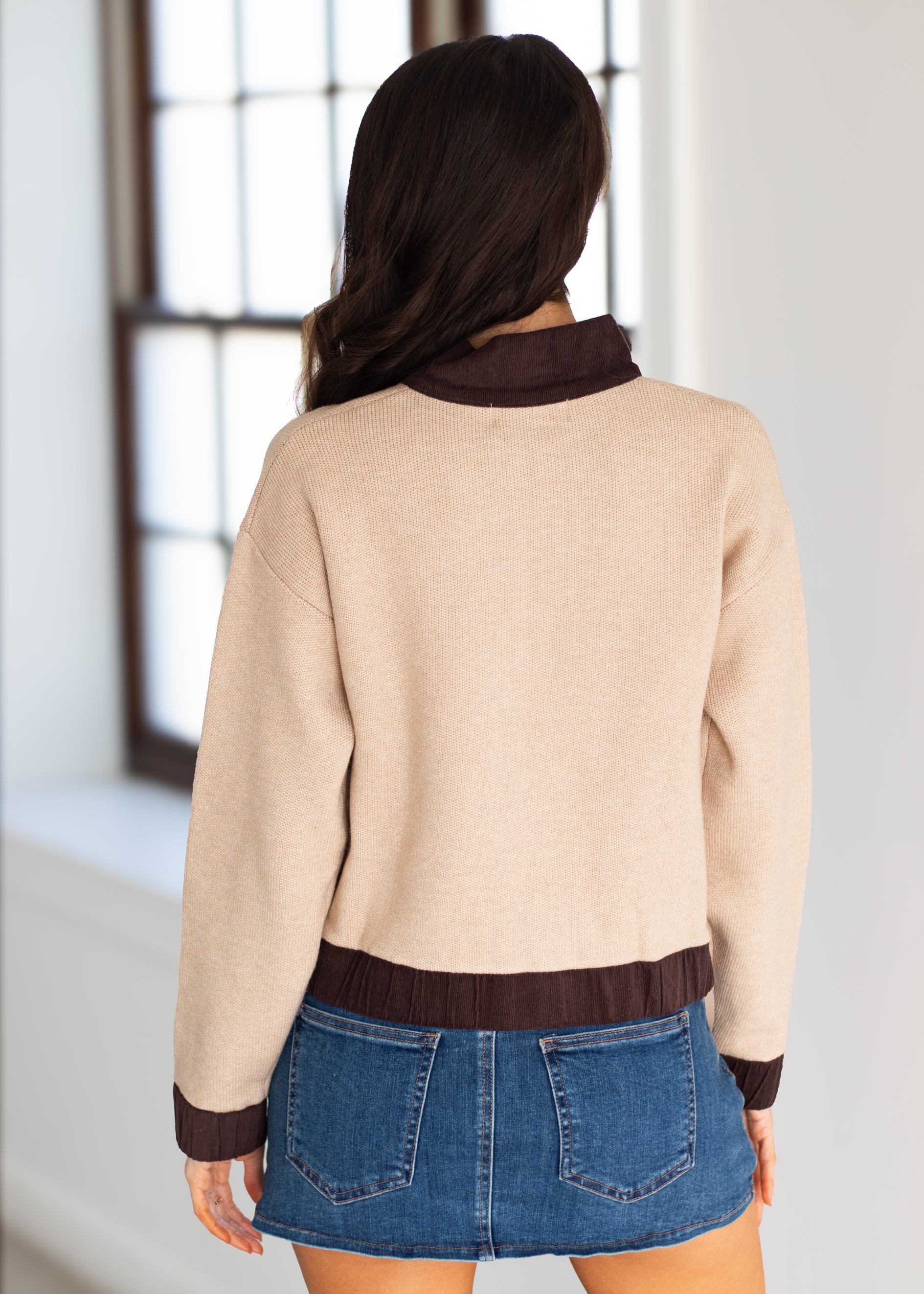 A woman shown from behind wearing a beige knit sweater with dark brown ribbed trim at the collar, cuffs, and hem, paired with a blue denim mini skirt in a bright indoor setting.