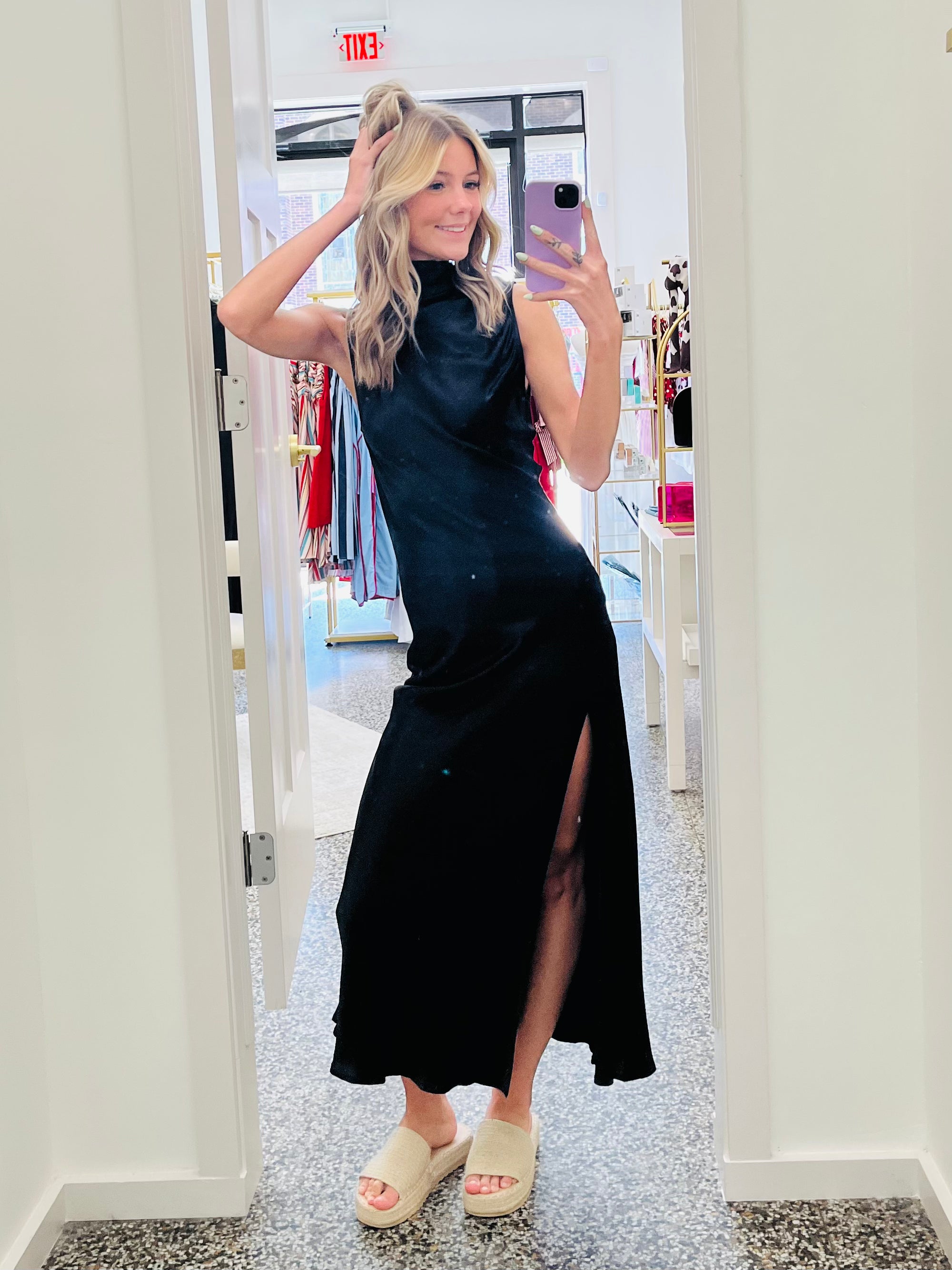 Woman taking a mirror selfie wearing a black dress with a slit in a store.
