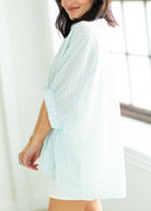 Woman wearing a light blue textured shirt over a white blouse with white shorts on a beige background

