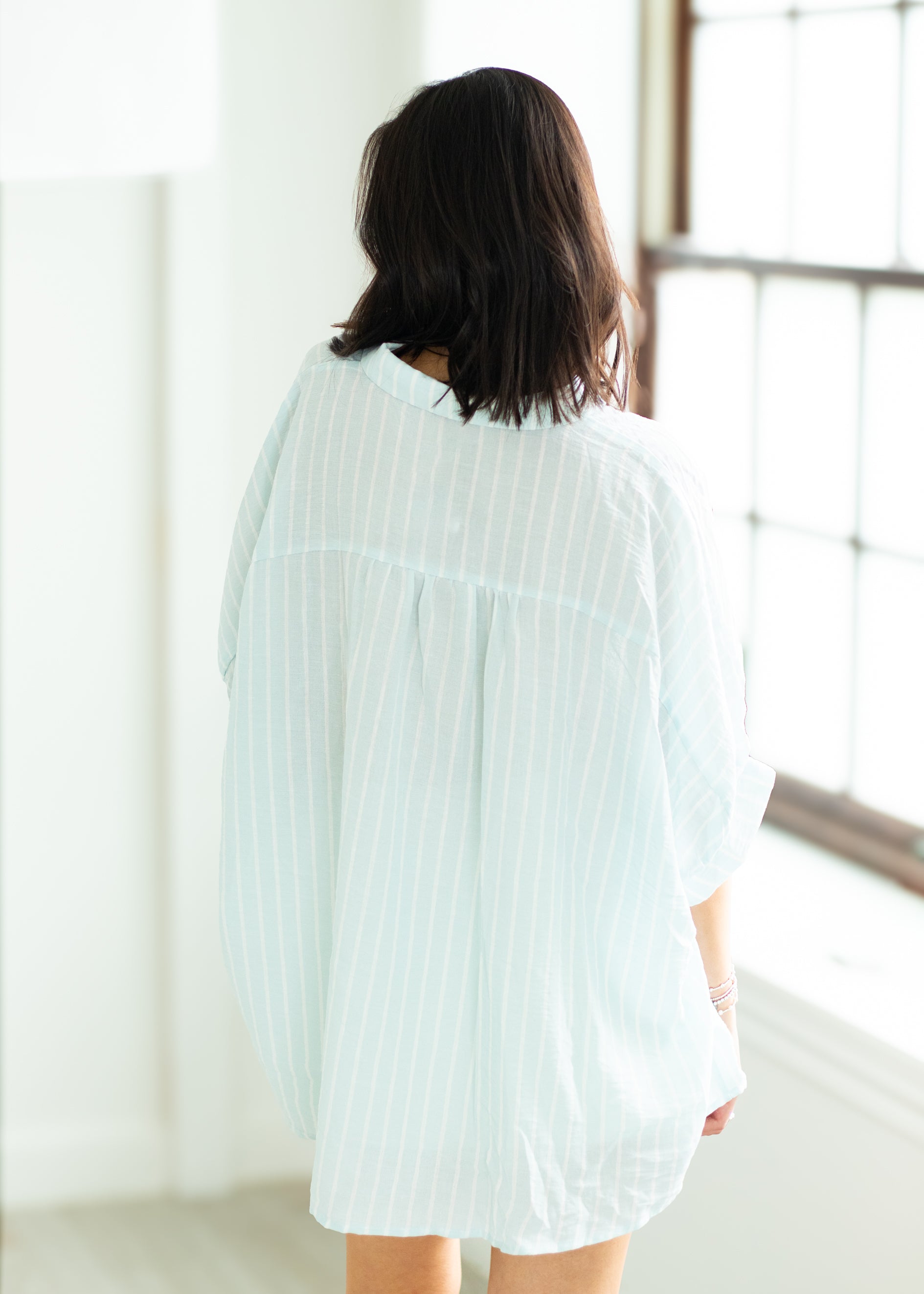 Woman wearing a light blue textured shirt over a white blouse with white shorts on a beige background

