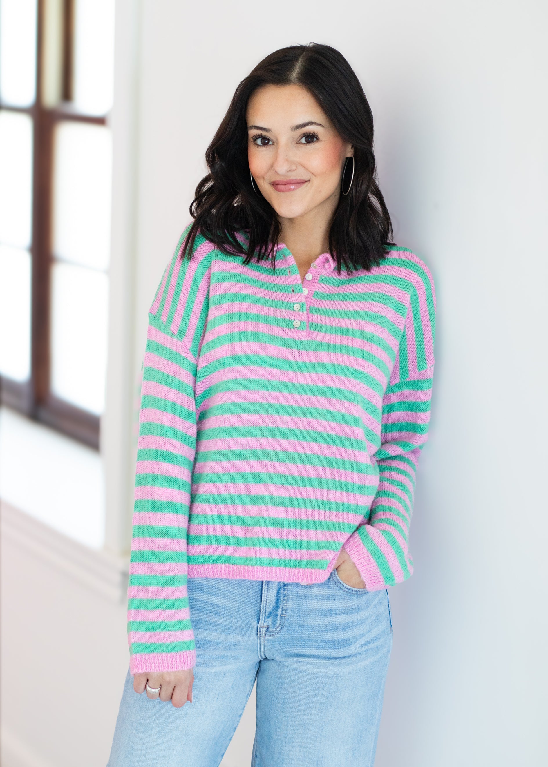 Woman wearing a pink and green striped sweater with blue jeans against a white wall.