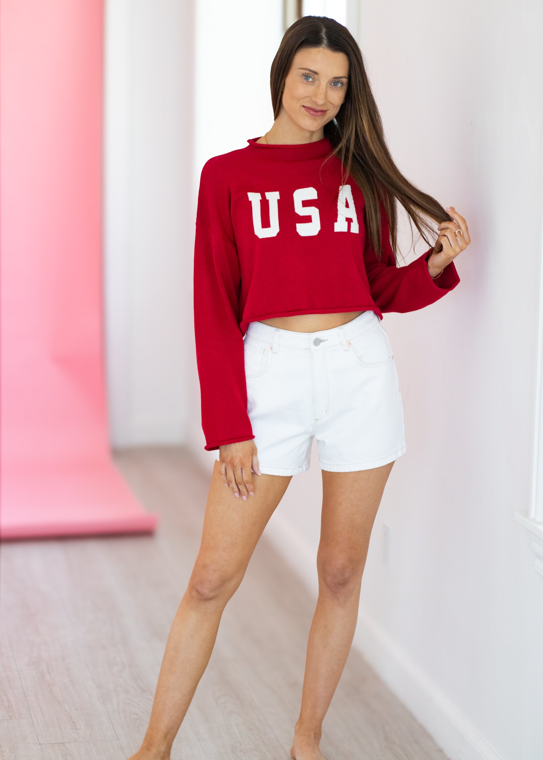 Woman wearing a red 'USA' sweatshirt and white shorts in a minimalistic setting.