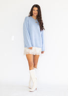 A woman modeling a light blue oversized knit sweater dress with a white lace hem, paired with white knee-high boots, against a plain white studio background.