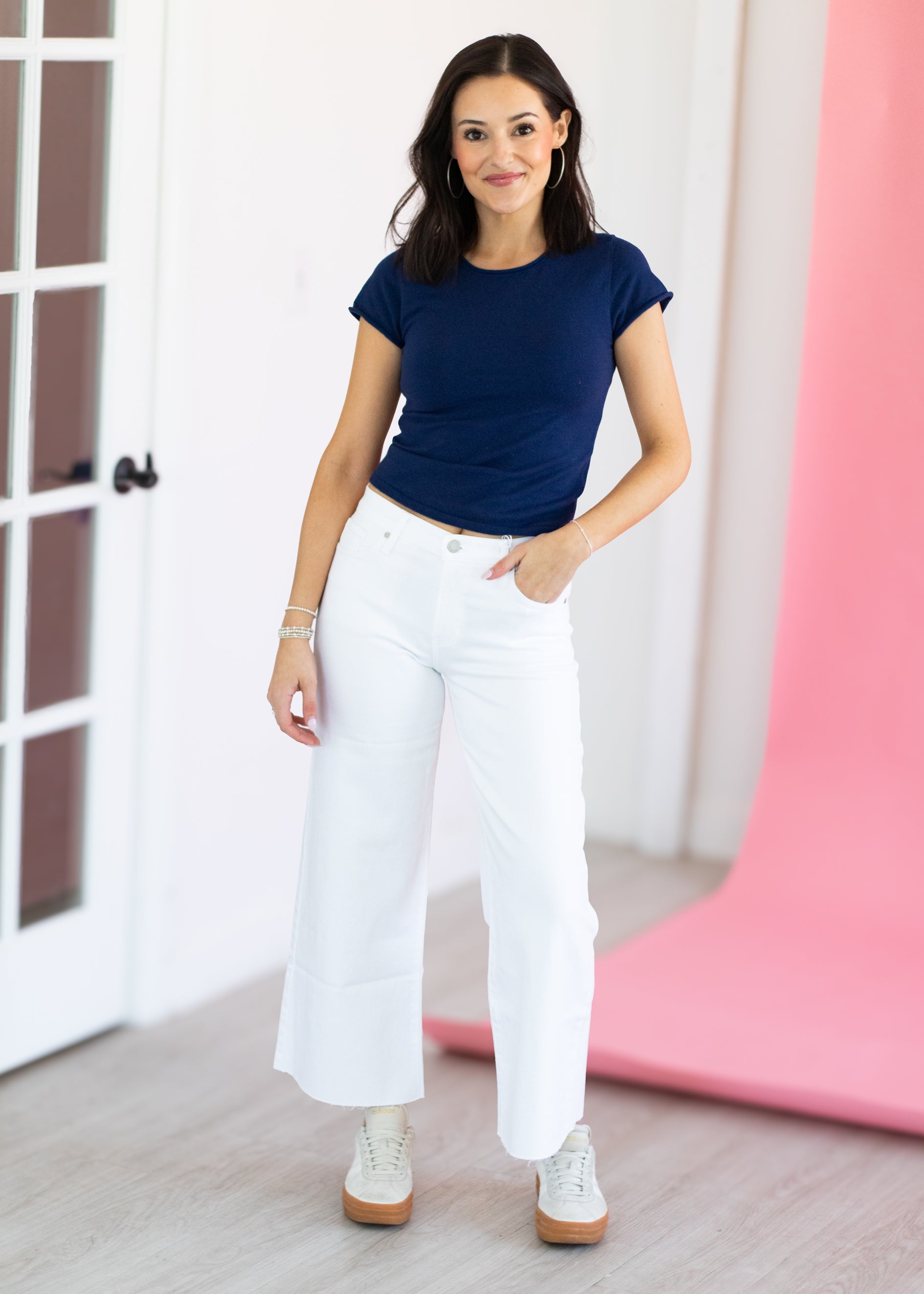 Woman wearing a navy blue top and white pants standing in a room with a pink curtain.