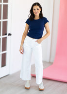 Woman wearing a navy blue top and white pants standing in a room with a pink curtain.