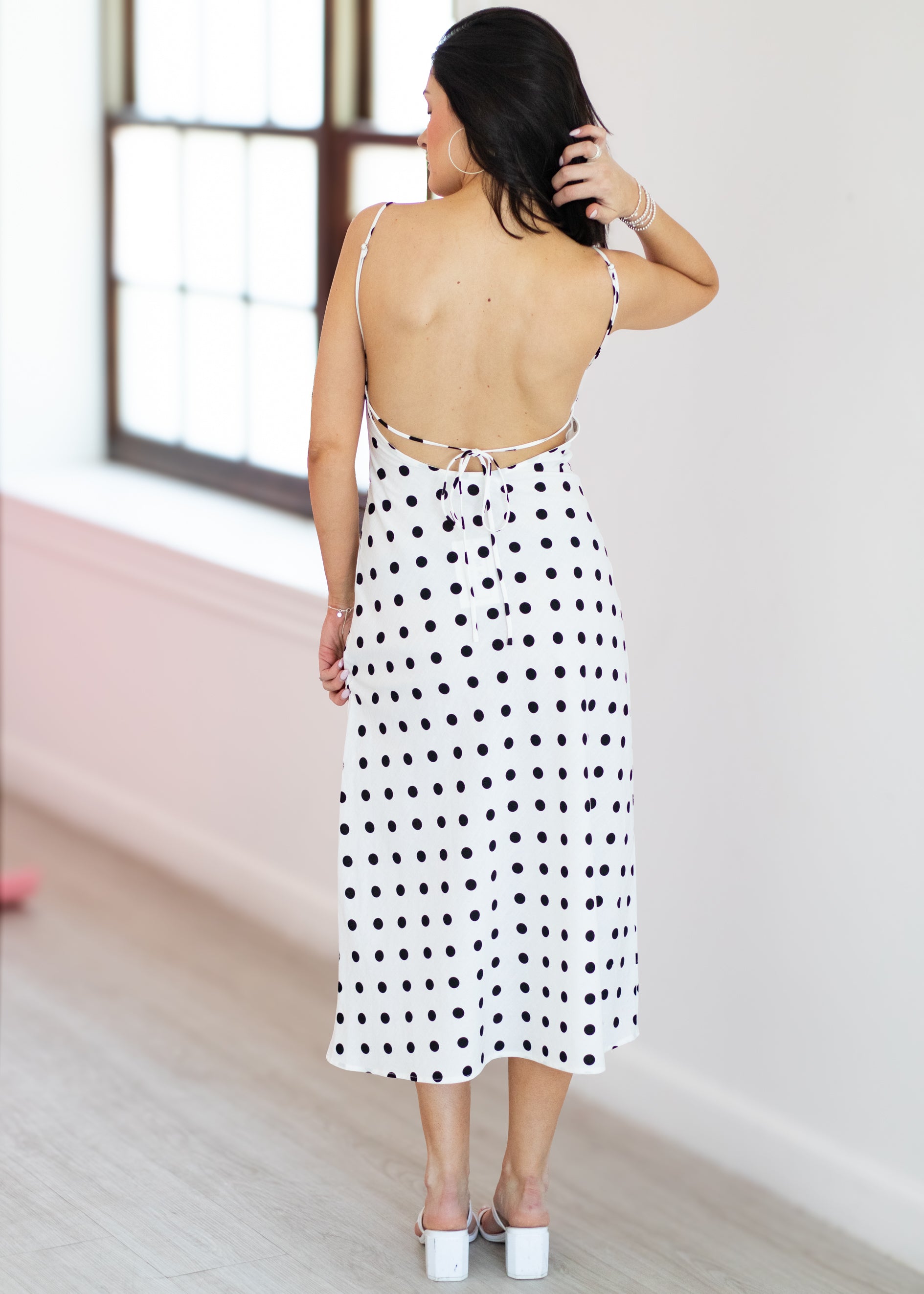 Woman wearing a polka dot dress with a backless design, standing in a room with a window.