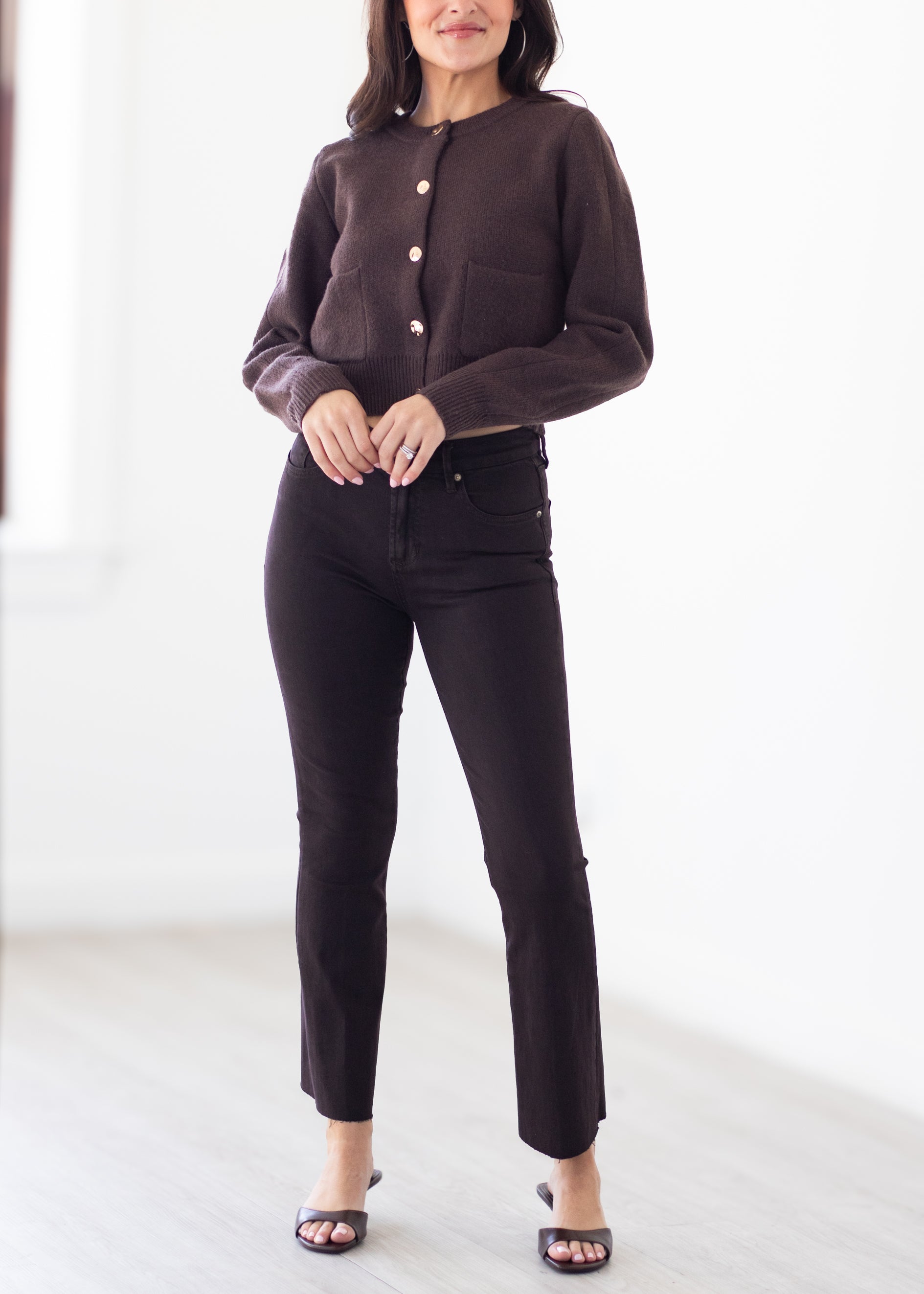 A woman modeling a dark brown button-front cardigan with a fuzzy texture and dark brown slim-fit jeans, standing in a bright, minimalist room.