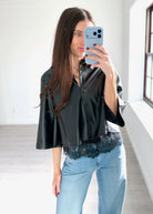 A woman takes a mirror selfie in a bright room, wearing a black faux-leather cropped button-up shirt with wide three-quarter sleeves and a black lace hem, paired with light blue jeans.