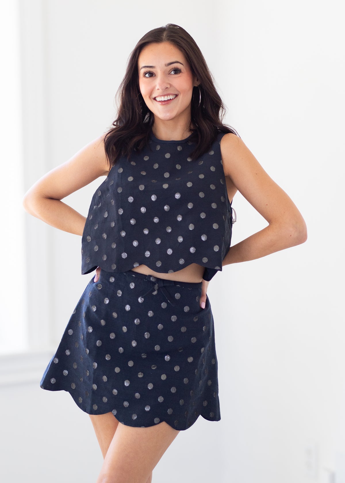 A woman modeling a matching sleeveless crop top and flared mini skirt set in navy blue with a silver polka dot pattern, set against a bright white indoor background.