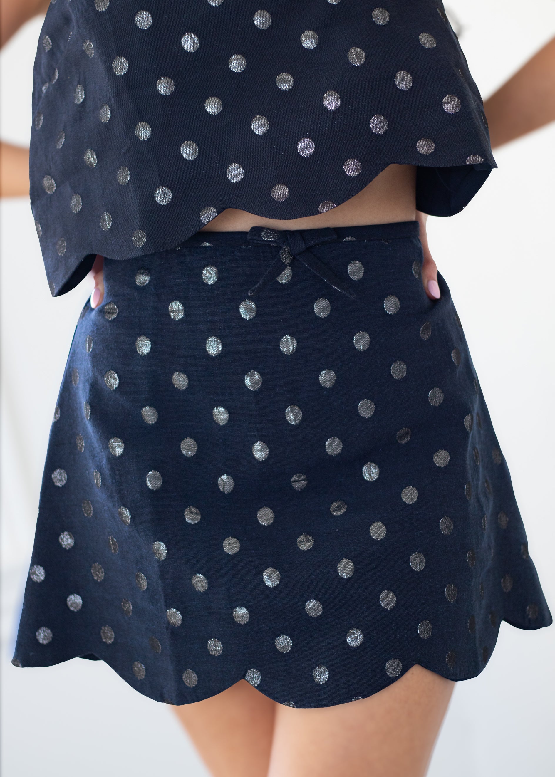 A close-up of a person wearing a matching navy blue two-piece set with metallic silver polka dots, featuring a scalloped hem and a tie detail at the waist.