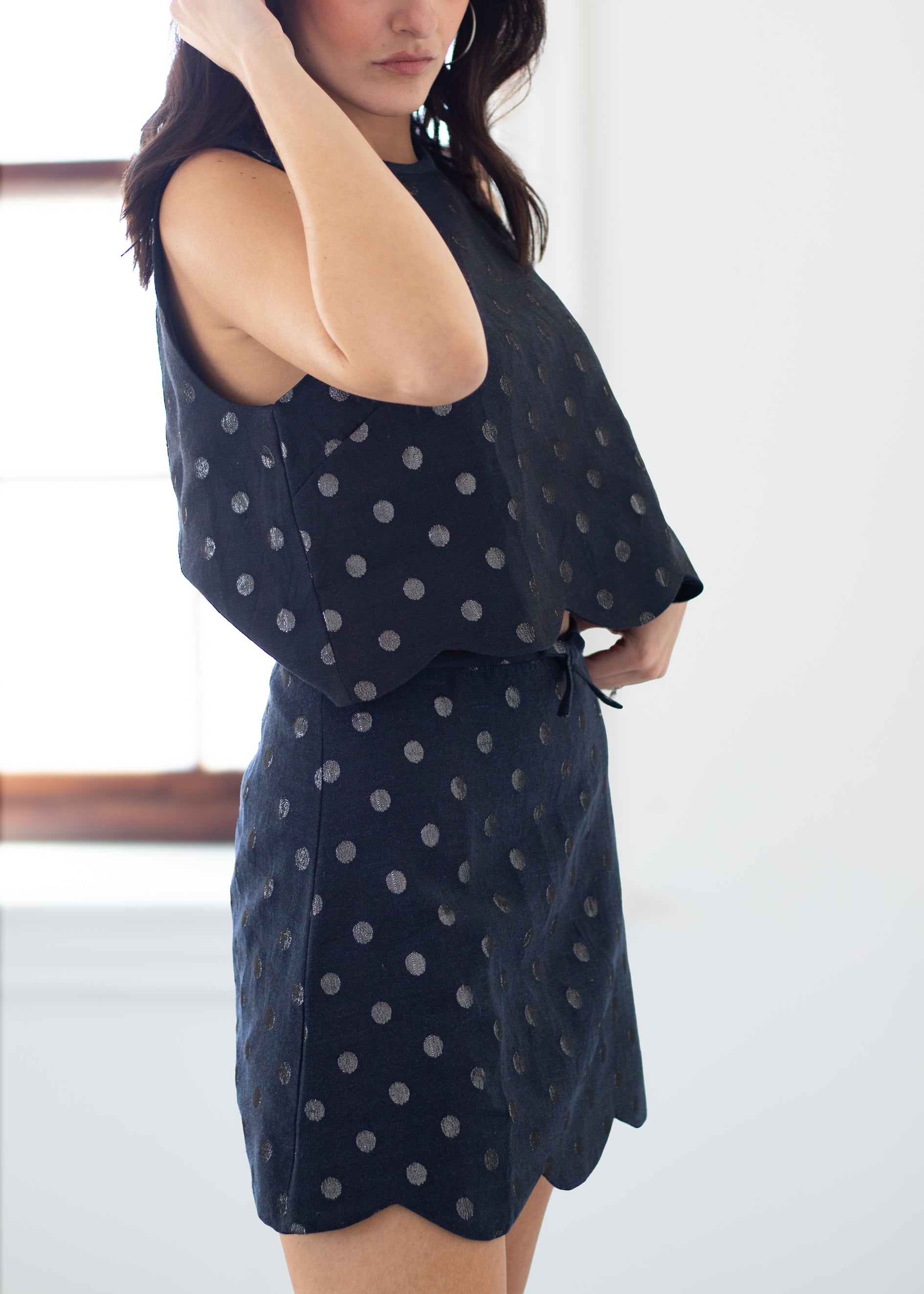 A woman wearing a sleeveless dark navy dress with a silver polka dot pattern, standing in a brightly lit room.