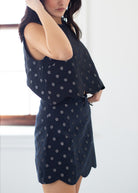 A woman wearing a sleeveless dark navy dress with a silver polka dot pattern, standing in a brightly lit room.